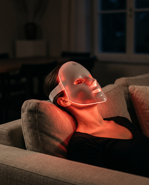 Woman reclining on a sofa wearing the Amora Glow Mask, lit red in a dim room